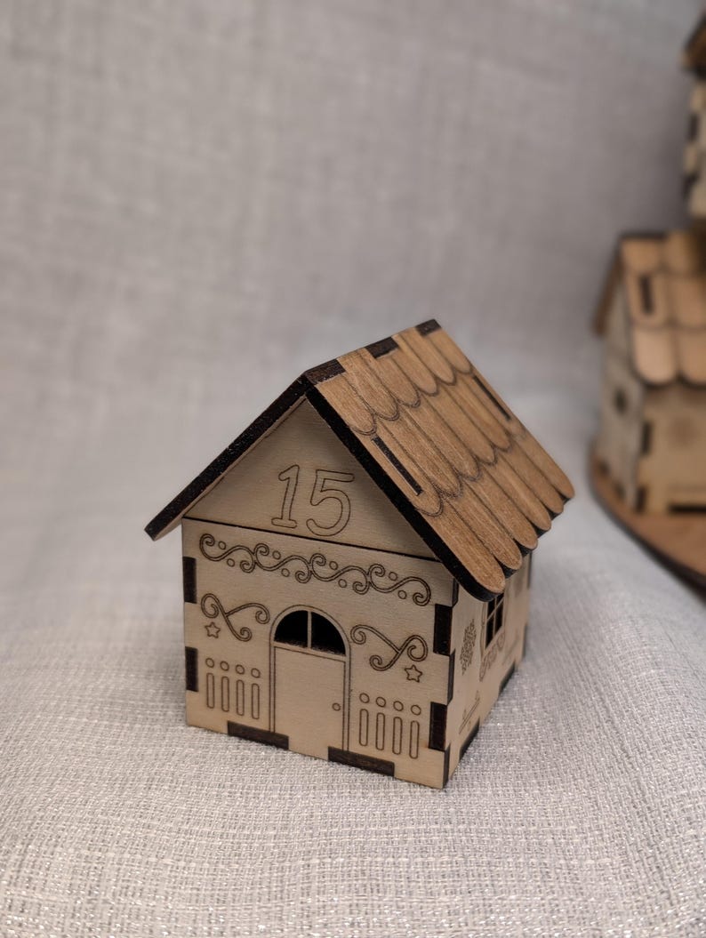 May include: A miniature wooden house with a gabled roof and decorative cutouts. The number "15" is displayed above the arched doorway. The house is made of light-coloured wood with a darker brown roof, and is likely a decorative item.