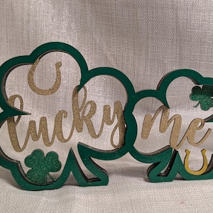 May include: A green wooden sign with the words "lucky me" in gold glitter. The sign is shaped like two overlapping shamrocks, with a horseshoe and shamrock details.