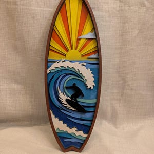 May include: A wooden surfboard-shaped wall art piece. The artwork depicts a surfer riding a wave with a sunset in the background. The sun is yellow and orange, and the wave is blue and white. The frame is brown.