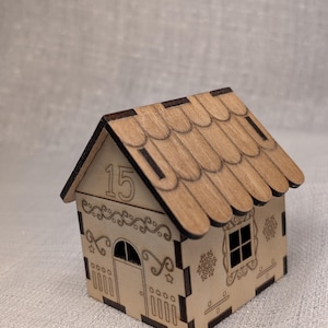 May include: A miniature wooden house with a scalloped roof and decorative cut-out patterns. The house has a door, window, and snowflake designs. The number "15" is displayed on the front. The house is made of light-coloured wood.
