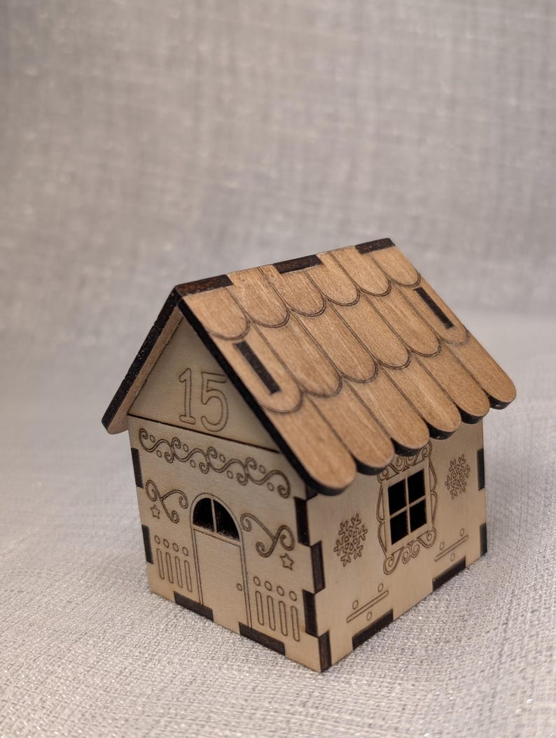 May include: A miniature wooden house with a scalloped roof and decorative cut-out patterns. The house has a door, window, and snowflake designs. The number "15" is displayed on the front. The house is made of light-coloured wood.