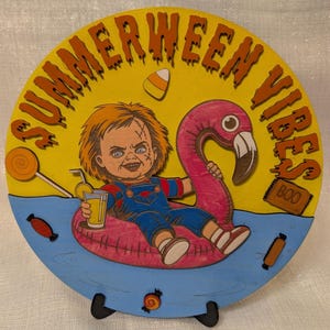 May include: A round wooden sign with the words "SUMMERWEEN VIBES" in orange lettering. It features a cartoon character in blue overalls, floating on a pink flamingo pool float, holding a drink. Candy corn and other candies are also depicted.