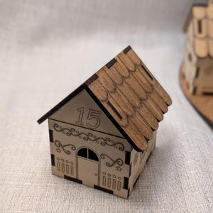 May include: A small, wooden house-shaped box with a brown shingled roof. The box is light brown with decorative cutouts and the number "15" on the front. The house has a door and window cut out of the front.