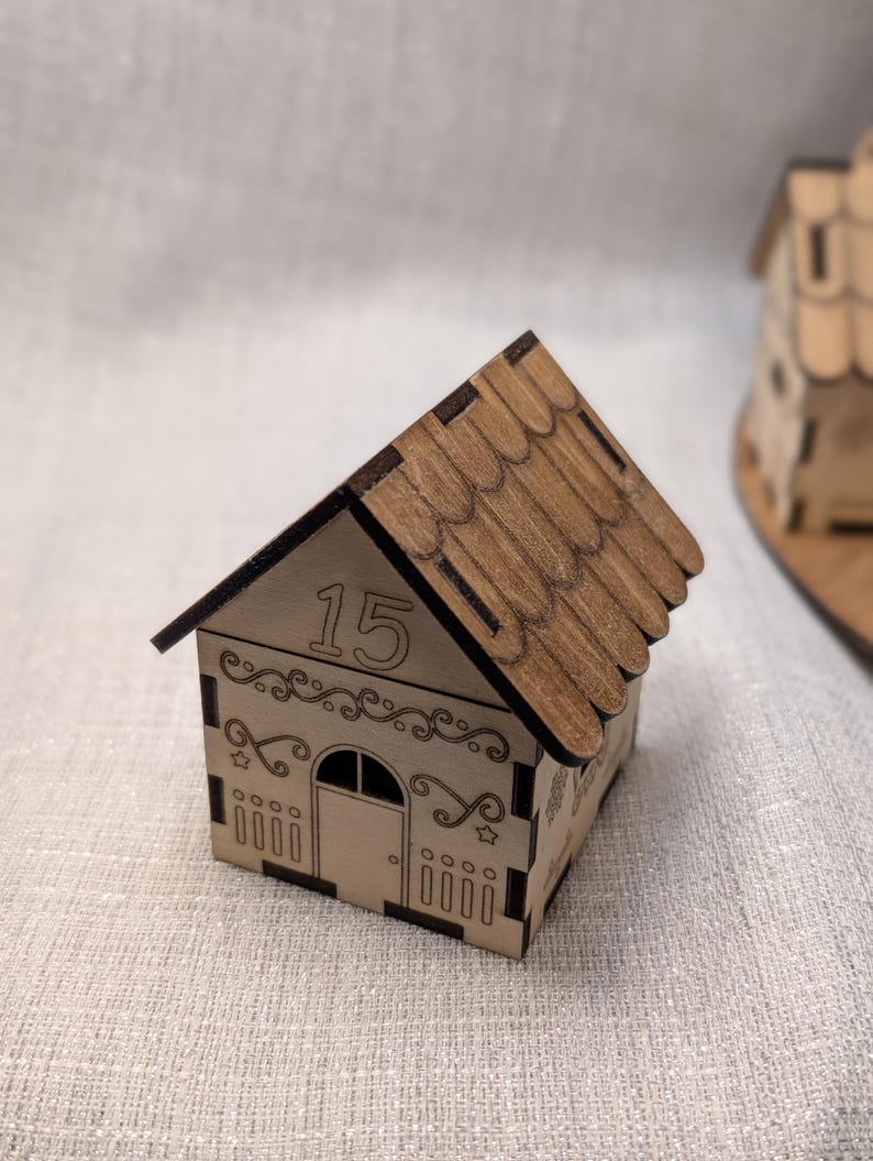 May include: A small, wooden house-shaped box with a brown shingled roof. The box is light brown with decorative cutouts and the number "15" on the front. The house has a door and window cut out of the front.