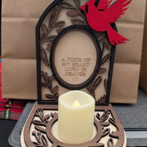 May include: A wooden memorial candle holder featuring a red cardinal and the text "A PIECE OF MY HEART LIVES IN HEAVEN." The candle holder has a floral design and holds a cream-coloured LED candle.