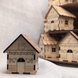 May include: A collection of small wooden houses with detailed cut-out designs. The house in the front displays the number 15 on its roof. Other houses are visible, suggesting a village or advent calendar theme. The houses are made of light-colored wood.
