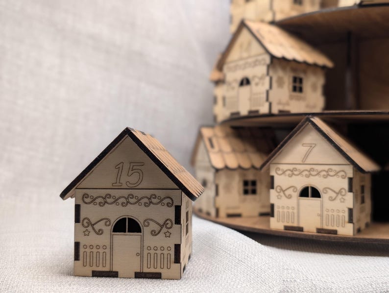 May include: A collection of small wooden houses with detailed cut-out designs. The house in the front displays the number 15 on its roof. Other houses are visible, suggesting a village or advent calendar theme. The houses are made of light-colored wood.