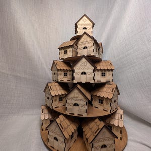 May include: A wooden advent calendar in the shape of a tiered village. Each house is laser-cut with detailed doors, windows, and patterns. Numbers are etched on the houses. The calendar is made of light brown wood.