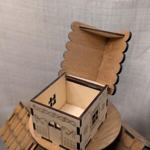 May include: A small, open wooden house-shaped box with a hinged, scalloped lid. The box features etched details of windows, a door, and decorative scrollwork. The box is sitting on a wooden platform, with other similar boxes in the background.