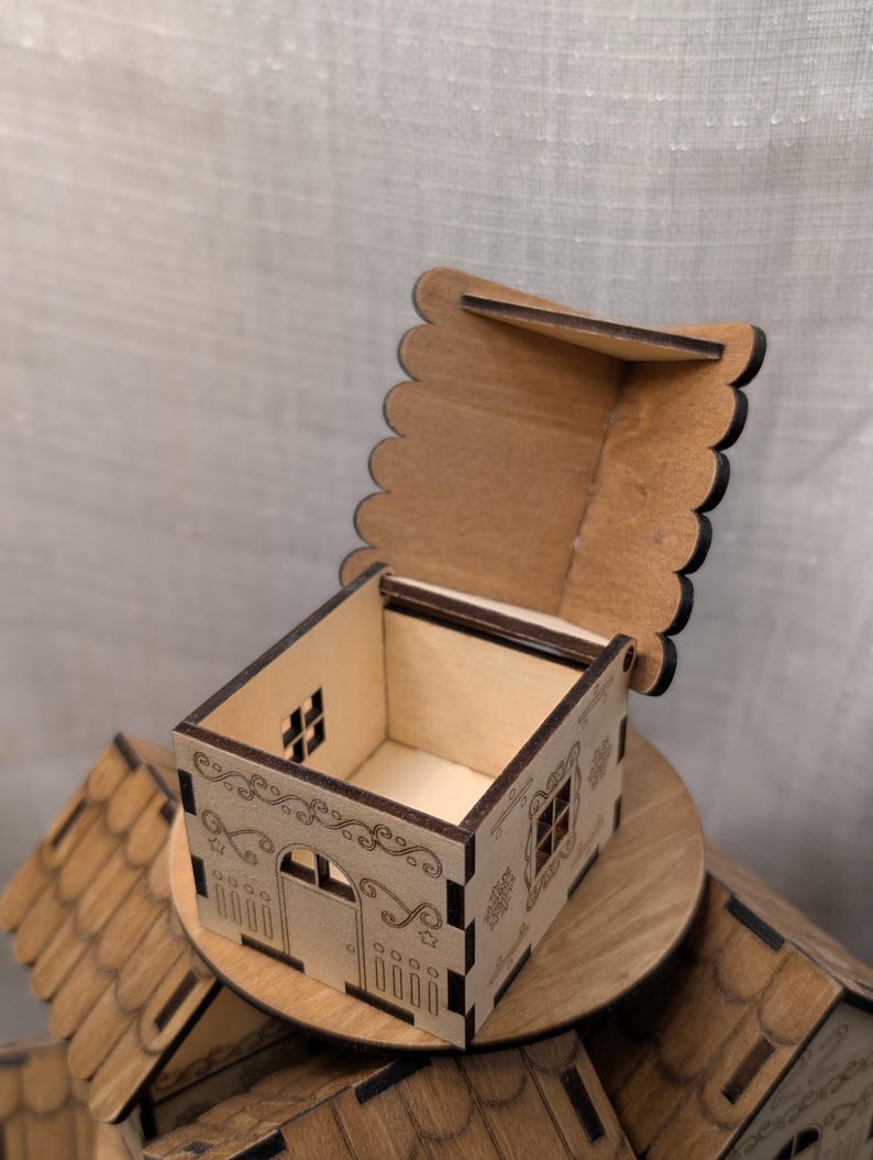 May include: A small, open wooden house-shaped box with a hinged, scalloped lid. The box features etched details of windows, a door, and decorative scrollwork. The box is sitting on a wooden platform, with other similar boxes in the background.