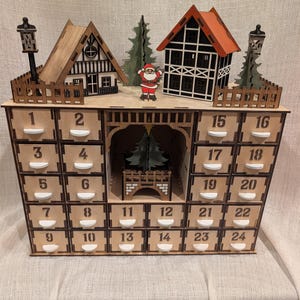 May include: A wooden advent calendar featuring twenty-four numbered drawers, a central archway with a tree, and a top display of two houses, two street lamps, two trees, and a Santa Claus figure. The structure is made of light-coloured wood.