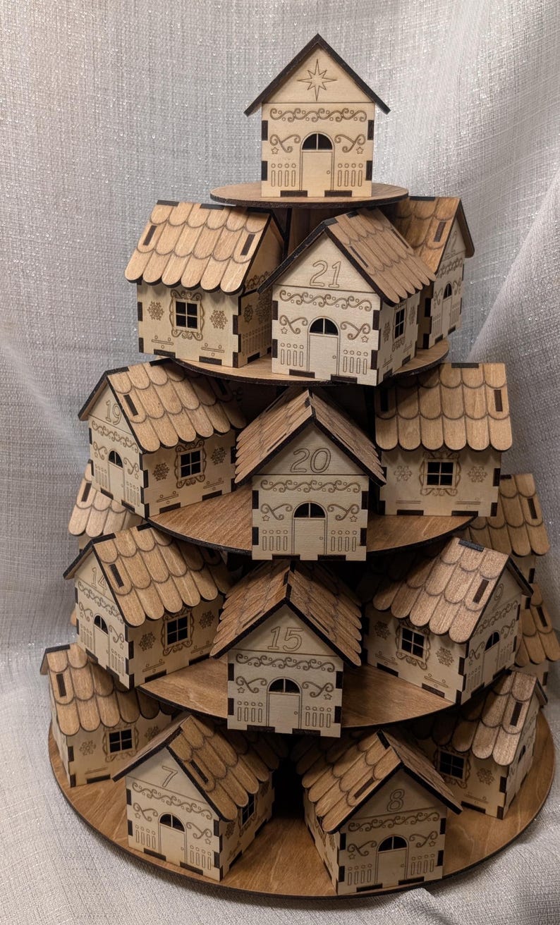 May include: A tiered wooden display of small, laser-cut houses, each with a number from 1 to 21. The houses have detailed roofs, doors, and windows. The display is made of light brown wood and resembles a Christmas tree.