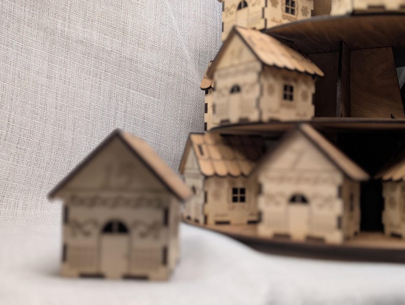May include: A display of miniature wooden houses, each with intricate cut-out details. The houses feature pitched roofs, arched doorways, and small windows. They are arranged on tiered platforms, forming a small village. The wood has a natural, light brown colour.