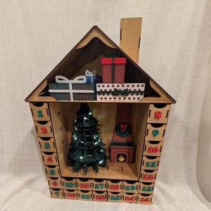 May include: A wooden house-shaped advent calendar featuring twenty-four small drawers numbered one to twenty-four in red and teal. The interior displays a dark green three-dimensional Christmas tree with lights and a brick fireplace cutout.