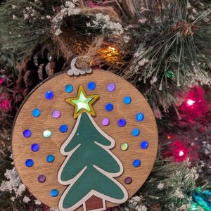 May include: Wooden Christmas tree decoration. The circular ornament features a green Christmas tree with a gold star. Blue and purple iridescent dots are scattered around the tree. It hangs from a jute string.