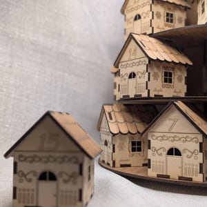 May include: Wooden miniature houses with decorative cutouts and numbers, arranged on tiered shelves. The houses have detailed roof designs, windows, and doors. The numbers 7, 14, and 15 are visible on the houses. The houses are made of light-coloured wood.