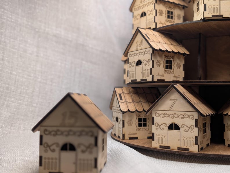 May include: Wooden miniature houses with decorative cutouts and numbers, arranged on tiered shelves. The houses have detailed roof designs, windows, and doors. The numbers 7, 14, and 15 are visible on the houses. The houses are made of light-coloured wood.