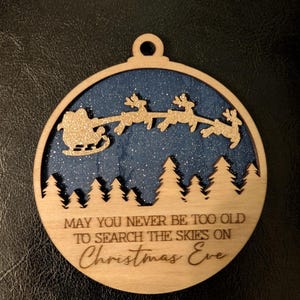 May include: Circular wooden Christmas ornament. The design features Santa Claus in a sleigh pulled by reindeer, set against a starry blue backdrop. The text reads "May you never be too old to search the skies on Christmas Eve."
