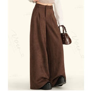 May include: High-waisted wide-leg trousers in a solid dark brown colour. These trousers feature a structured waistband with a single button closure, a front zip fly, and subtle pleats at the waistline, falling into a loose, floor-length silhouette.