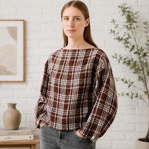 May include: A long-sleeved blouse featuring a brown and white plaid pattern with a wide boat neckline and a loose, boxy fit. The garment has a straight hemline and a single visible button on the cuff of each sleeve, paired with grey denim jeans.