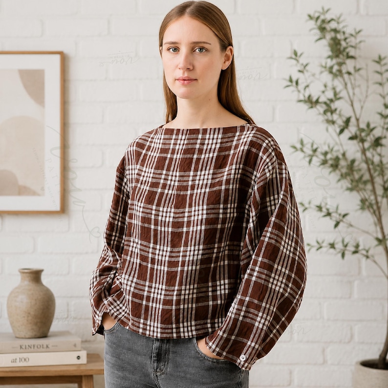 May include: A long-sleeved blouse featuring a brown and white plaid pattern with a wide boat neckline and a loose, boxy fit. The garment has a straight hemline and a single visible button on the cuff of each sleeve, paired with grey denim jeans.