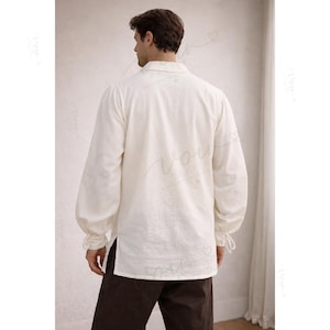 May include: A long-sleeved, off-white shirt featuring a fold-over collar, a button-down front, and gathered cuffs with ties at the wrists. The shirt has a straight hem with side slits and is paired with dark brown trousers.