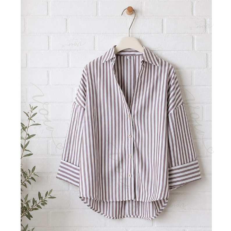 May include: A button-down shirt featuring a vertical striped pattern in muted brown and white. The shirt has a classic collar, three-quarter length sleeves with wide cuffs, and a slightly curved hemline, hanging on a light-colored plastic hanger.