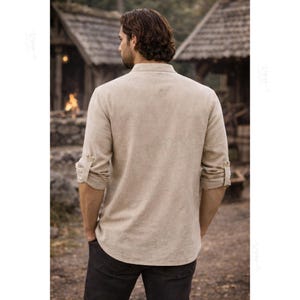 May include: A beige long-sleeved shirt with a mandarin collar and button-tab sleeves, shown from the back. The shirt features a textured fabric, a straight hemline, and a buttoned placket detail on the sleeves, paired with dark trousers.