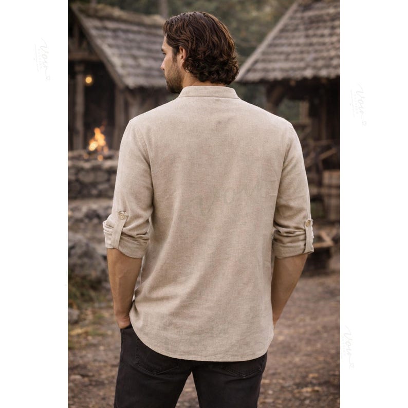May include: A beige long-sleeved shirt with a mandarin collar and button-tab sleeves, shown from the back. The shirt features a textured fabric, a straight hemline, and a buttoned placket detail on the sleeves, paired with dark trousers.