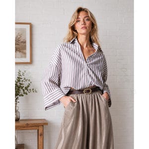 May include: A long-sleeved button-down shirt featuring vertical white and grey stripes, paired with high-waisted metallic taupe trousers. The trousers are cinched at the waist with a dark brown leather belt that has a gold-toned oval buckle.