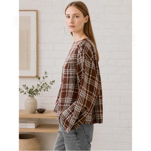 May include: A long-sleeved shirt featuring a brown and white plaid pattern with a round neckline and a relaxed fit. The shirt is paired with grey denim jeans. The fabric has a visible textured weave, and the sleeves are loose and straight.
