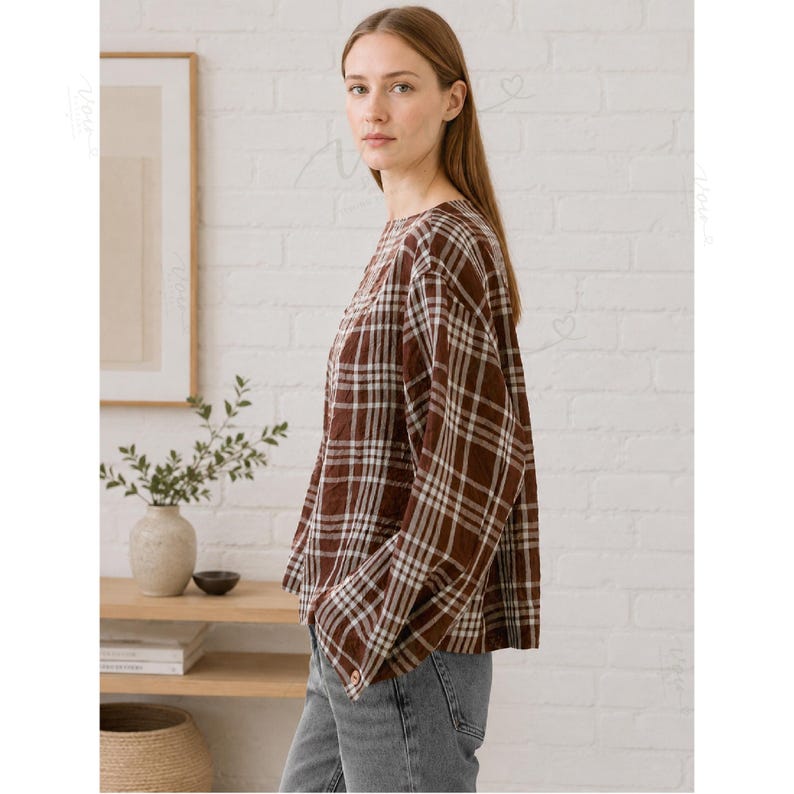 May include: A long-sleeved shirt featuring a brown and white plaid pattern with a round neckline and a relaxed fit. The shirt is paired with grey denim jeans. The fabric has a visible textured weave, and the sleeves are loose and straight.