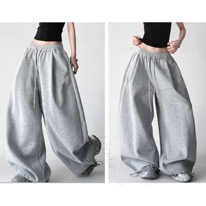 May include: Light grey sweatpants featuring a high-waisted design with an elastic waistband and a drawstring closure. These wide-leg trousers have a loose, relaxed fit that drapes down to the ankles, providing a voluminous silhouette.