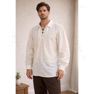 May include: An off-white long-sleeve shirt featuring a pointed collar and a front lace-up placket with matching fabric ties. The garment has full sleeves with gathered cuffs and a straight hemline, constructed from a textured, woven fabric.