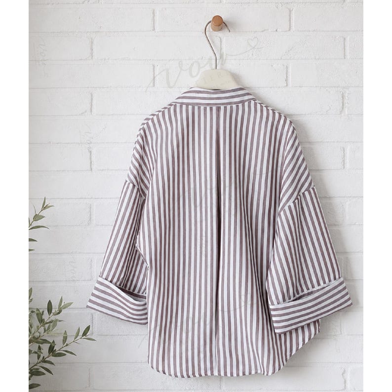 May include: A button-down shirt featuring a vertical striped pattern in dark brown and white. The shirt has a classic collar, wide three-quarter length sleeves with folded cuffs, and a loose, relaxed fit, displayed hanging on a white hanger.
