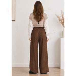 May include: A pair of dark brown wide-leg trousers paired with a white long-sleeved shirt and a beige sleeveless knit vest. The trousers feature a high-waisted design with belt loops and a smooth, matte texture, worn with black shoes.