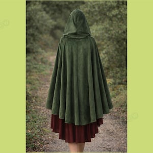 May include: A dark green hooded cloak with a wide, flowing design. The cloak is made of a soft, velvety material and is worn over a burgundy skirt. The person is standing on a path in a forest setting.