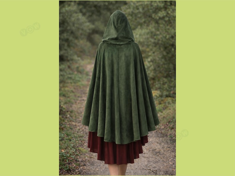 May include: A dark green hooded cloak with a wide, flowing design. The cloak is made of a soft, velvety material and is worn over a burgundy skirt. The person is standing on a path in a forest setting.