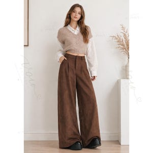 May include: A beige V-neck knit sweater vest layered over a white long-sleeve button-down shirt, paired with high-waisted brown wide-leg trousers featuring a front button closure and black platform shoes.