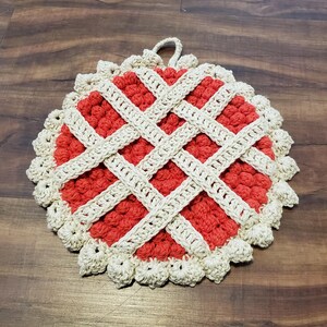 May include: A handmade, round crochet hot pad. The centre is a textured red, overlaid with a lattice of cream-coloured yarn. The edge is scalloped, and a loop is attached for hanging. A charming kitchen accessory.