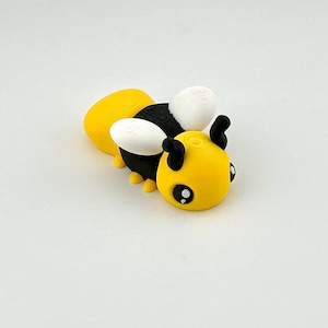 May include: A 3D-printed bee figurine in yellow, black, and white. The bee has large, cartoonish eyes and small antennae. The wings are white, and the body is yellow and black. The figurine is a cute, whimsical design.