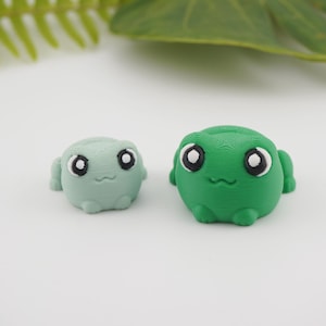 May include: Two small, round frog figurines. One is light green, the other is bright green. Both have large, white-rimmed eyes with black pupils and small, smiling mouths. The frogs have small arms and legs.