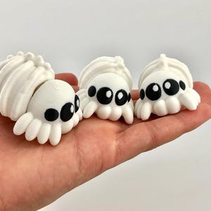 May include: Three white, 3D-printed spider figurines with black eyes and small, rounded legs. The spiders have a textured, shell-like back. The figurines are held in a hand against a white background.