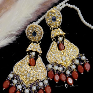 May include: Gold-toned, ornate earrings with a teardrop design. The earrings feature clear, sparkling stones, red-brown beads, and white pearls. The earrings are suspended from a white beaded chain, with a black background.