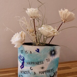 May include: A white ceramic vase with a black rim, decorated with blue watercolor-style designs and the words "IT IS A HAPPINESS TO WONDER, A HAPPINESS TO DREAM." The vase holds dried white flowers and grasses.