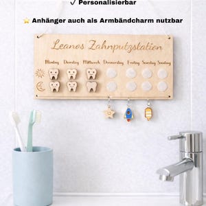 Könnte beinhalten: Zahnbürsten-Tafel aus Holz mit dem Text "Leanos Zahnputzstation" und den Wochentagen. Enthält zahnförmige und runde Aufkleber sowie Anhänger zum Aufhängen. Ein hellblauer Zahnbürstenhalter mit zwei Zahnbürsten befindet sich im Vordergrund.