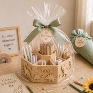 May include: A wooden hexagonal desk organiser featuring dinosaur cutouts and the name Theo, filled with coloured pencils, pens, and scissors. The item is wrapped in clear plastic with a sage green ribbon and a tag reading Zur Einschulung.