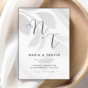 May include: A wedding invitation with a minimalist design. The invitation features the names "Nakia & Travis" in a simple font, along with the wedding date, time, and location. The invitation has a white background with a subtle leaf pattern.