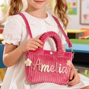 Peut inclure: Une jeune fille tient un sac à main en velours côtelé rose avec le nom "Amelia" brodé dessus. Le sac a une poignée supérieure et une bandoulière. Le sac est décoré d'accents floraux. La fille porte une robe blanche.