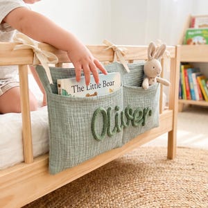 Könnte beinhalten: Ein Bett-Organizer aus salbeigrünem Musselin mit zwei Taschen, der mit cremefarbenen Bändern an einem Holzbettrahmen befestigt ist. Der Name Oliver ist in dunkelgrünem Garn auf die Vorderseite gestickt. Eine Tasche enthält ein Buch.